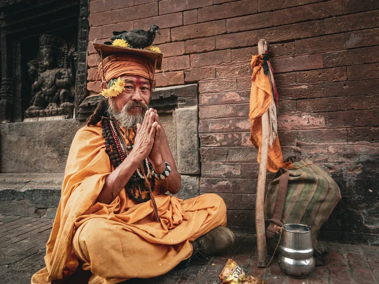 old yogi in orange clothes with a pigeon on his head is doing namaste pose while sitting in a temple with stick, bag and mug by his side | Picture by: Mehmet Turgut  Kirkgoz : https://www.pexels.com/de-de/foto/mann-kostum-verkleidung-religion-12226990/