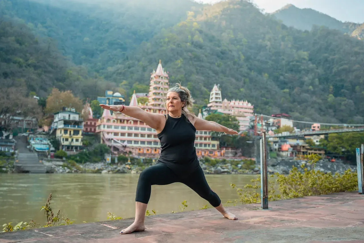 mature woman doing yoga warrior pose in a anchient river-side asian mountain city | Picture by Yoga Course  India von Pexels: https://www.pexels.com/de-de/foto/frau-wahrzeichen-stehen-fluss-20035456/